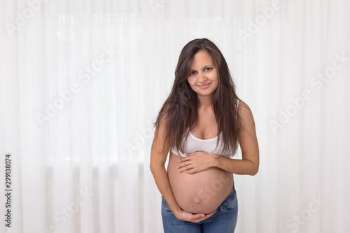 Pregnant woman in jeans and white top holds hands on belly on a white background. Pregnancy, maternity, preparation and expectation concept. Beautiful tender mood photo of pregnancy. Free Download Pregnant woman in jeans and white top holds hands on belly on a white background. Pregnancy, maternity, preparation and expectation concept. Beautiful tender mood photo of pregnancy. - GFXLoad