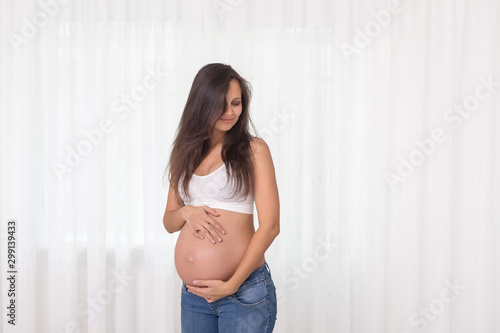 Pregnant woman in jeans and white top holds hands on belly on a white background. Pregnancy, maternity, preparation and expectation concept. Beautiful tender mood photo of pregnancy.