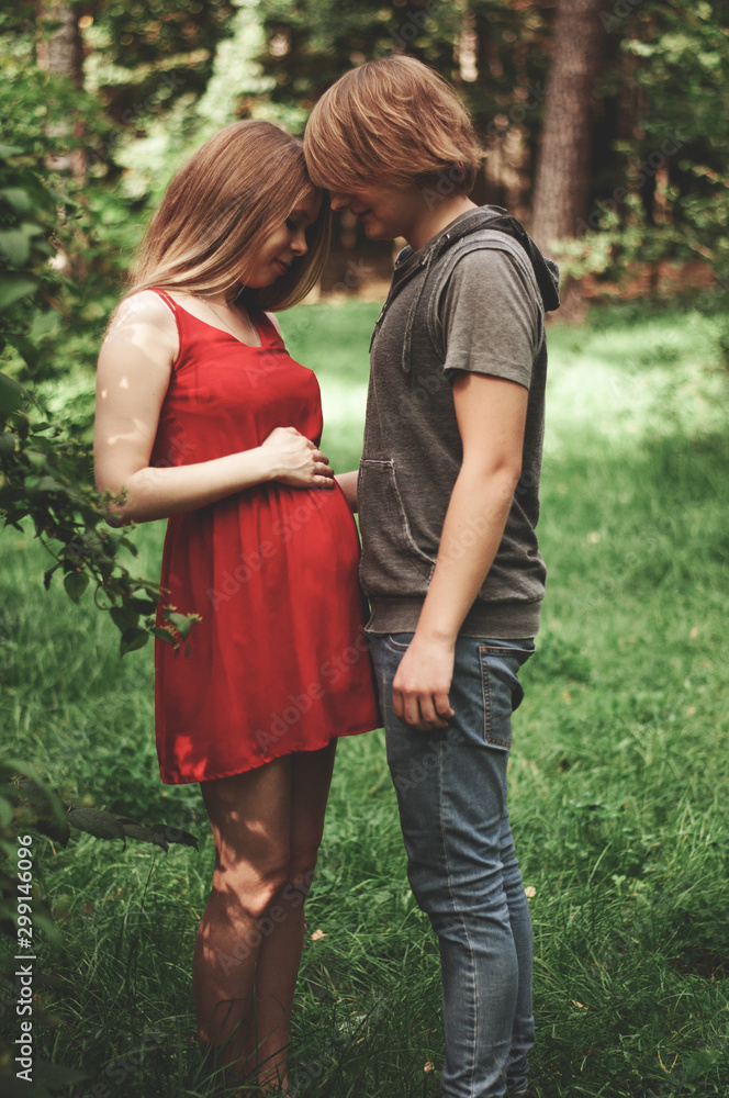 Fototapeta premium A happy married couple Husband hugs a beautiful adult pregnant wife. walks in a park. Second trimester. Holds his hands on her stomach. The benefits of walking during pregnancy, health care.