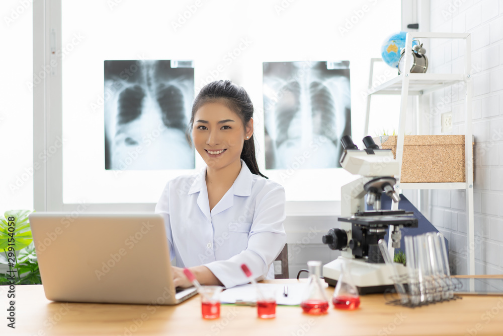 Asian female doctor working on laptop with a microscope for an exam at hospital giving patient convenience online service advice, smiling write a prescription , healthcare, preventing disease concept Asian female doctor working on laptop with a microscope for an exam at hospital giving patient convenience online service advice, smiling write a prescription , healthcare, preventing disease concept