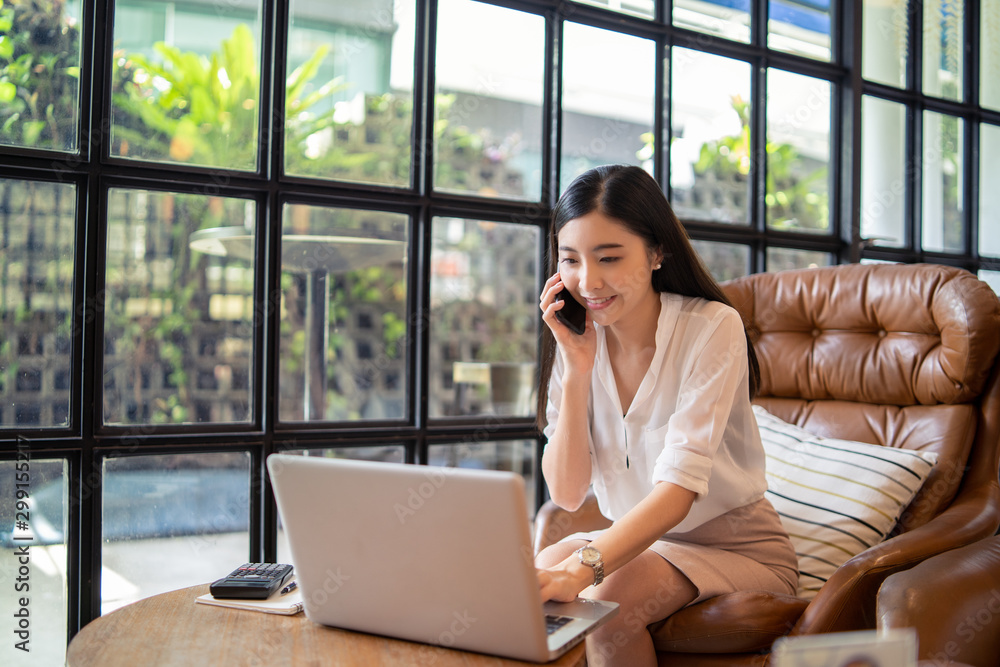 Smiling Business girl working in office with laptop,Happy Asian business woman using phone sitting on a chair at modern home studio.Concept of young people working mobile devices,contact to customer Smiling Business girl working in office with laptop,Happy Asian business woman using phone sitting on a chair at modern home studio.Concept of young people working mobile devices,contact to customer