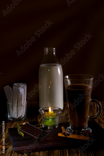 Glass of coffee on a wooden table with a bottle of milk, sugar bags, cologne and spices lit by candles.