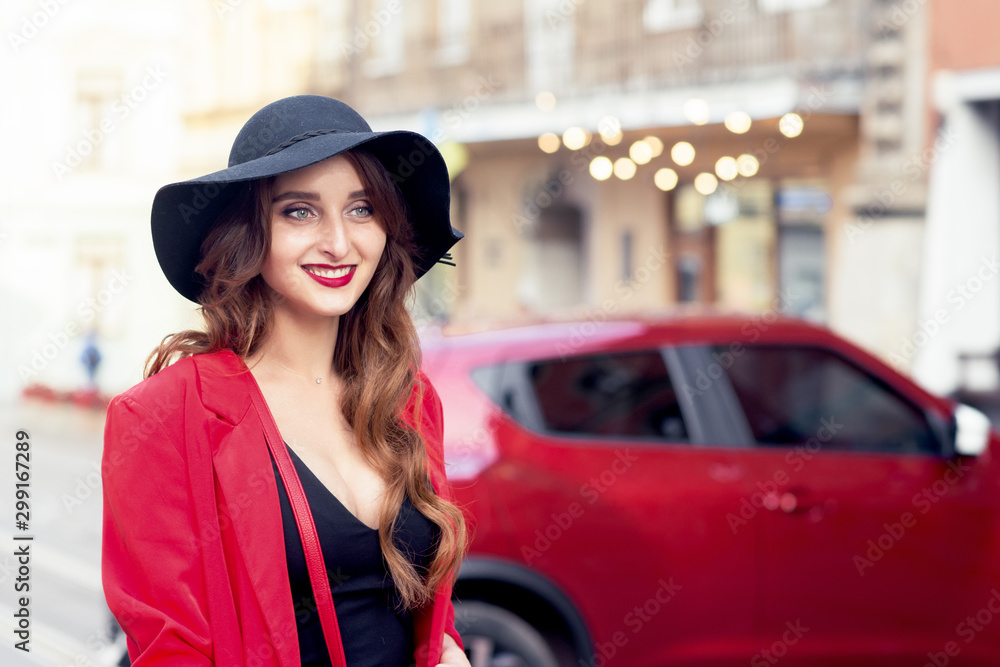 Beautiful caucasian smiling young woman is wearing black hat on the red car background on the city street.
