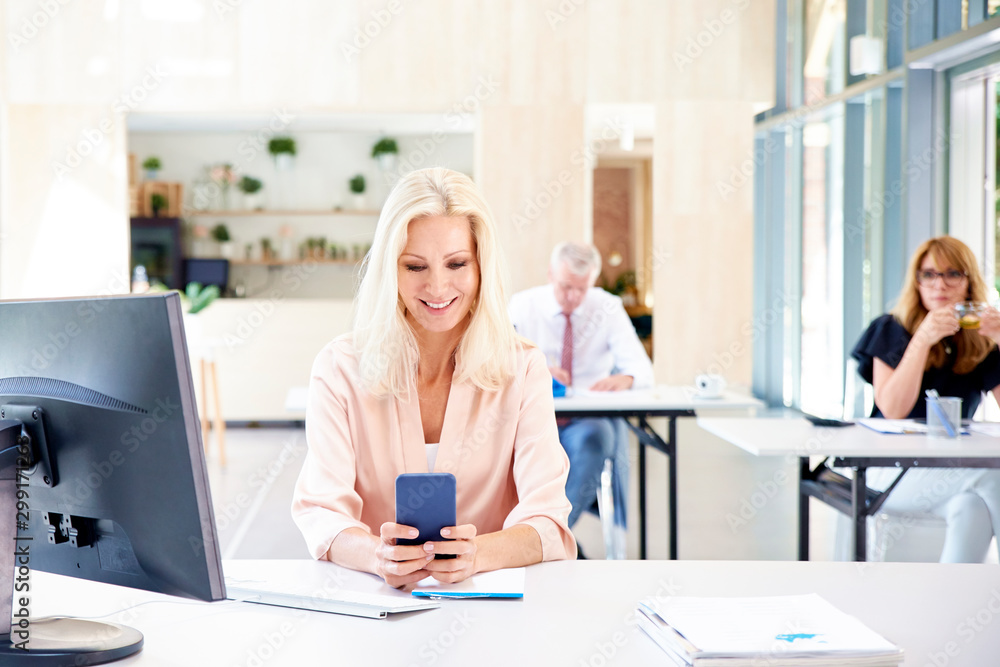 Shot of smiling blonde businesswoman text messaging while working on computer in the office. Shot of smiling blonde businesswoman text messaging while working on computer in the office.