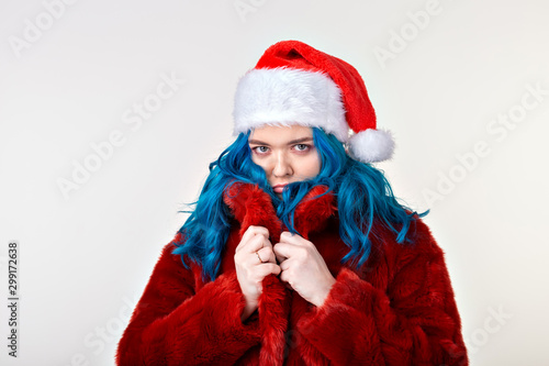 Funny caucasian woman in glamorous red fur coat and christmas santa hat posing on white background. Cheerful female model with blue hair having fun during photoshoot.