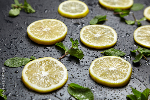 Lemon slices and green mint leaves on black background with water drops. Fresh tropical fruit, yellow citrus on dark stone table © mikeosphoto