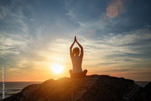 Yoga silhouette of woman meditating on the rocks near the ocean coastline during amazing sunset.