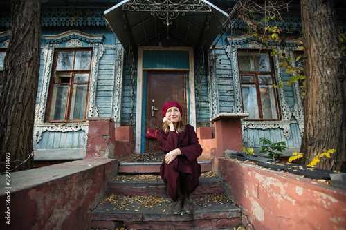 A woman in a burgundy coat and beret is waiting sitting on the porch of an old wooden house. © De Visu