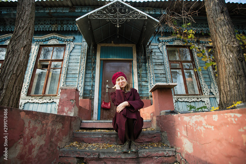 A woman in a burgundy coat and beret is patiently waiting sitting on the porch of an old wooden house. © De Visu