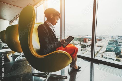 Side view of a dazzling young African-American woman entrepreneur sitting with a digital tablet on a yellow curved armchair indoors of a luxurious office room on a top floor of a business skyscraper