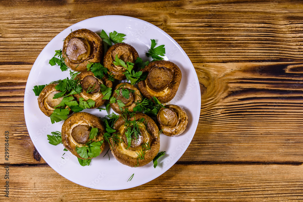 Plate with baked champignons, dill and parsley on a wooden table. Top view Plate with baked champignons, dill and parsley on a wooden table. Top view