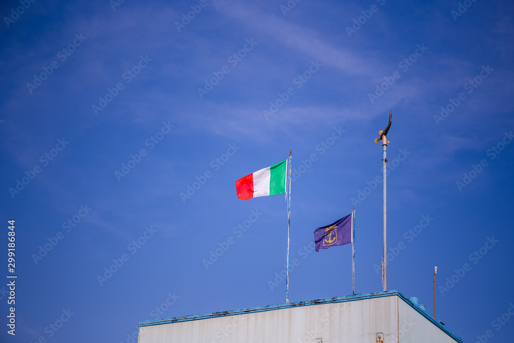 Flag of Italy and a blue flag with golden anchor on a building with blue sky in the background