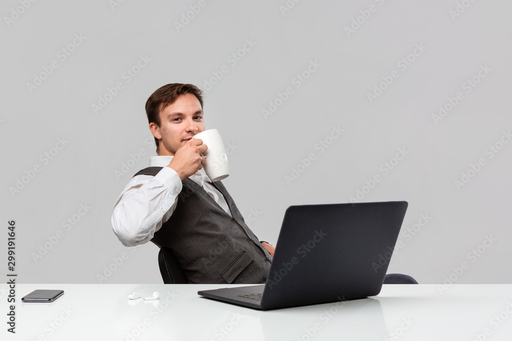 Businessman in a white t-shirt and grey vest drinking coffee sitting at the white table with laptop and smartphone. Businessman in a white t-shirt and grey vest drinking coffee sitting at the white table with laptop and smartphone.