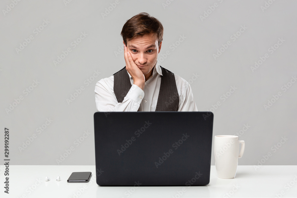 Office worker in a white t-shirt and grey vest is shocked checking information on a laptop sitting at the white table. Office worker in a white t-shirt and grey vest is shocked checking information on a laptop sitting at the white table.