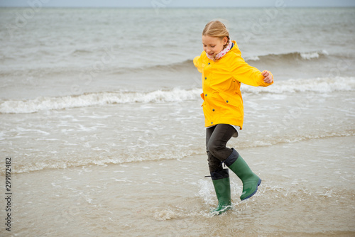 Cheerful little girl running on water of Baltic sea in rubber boots at windy weather
