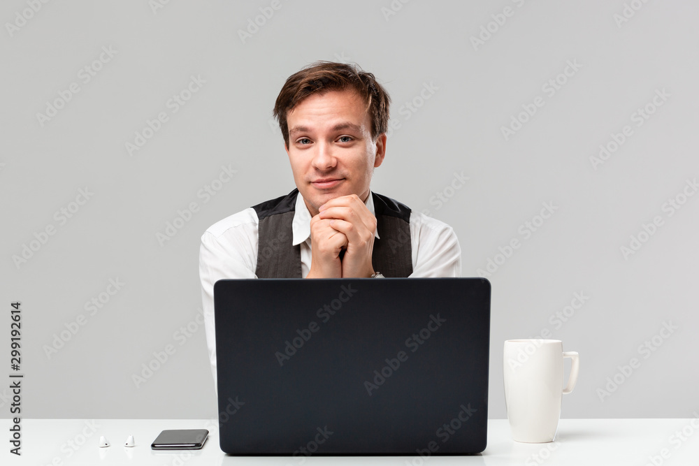 Businessman in a white t-shirt and grey vest working on the laptop in the office sitting at the white table with laptop and smartphone. Businessman in a white t-shirt and grey vest working on the laptop in the office sitting at the white table with laptop and smartphone.