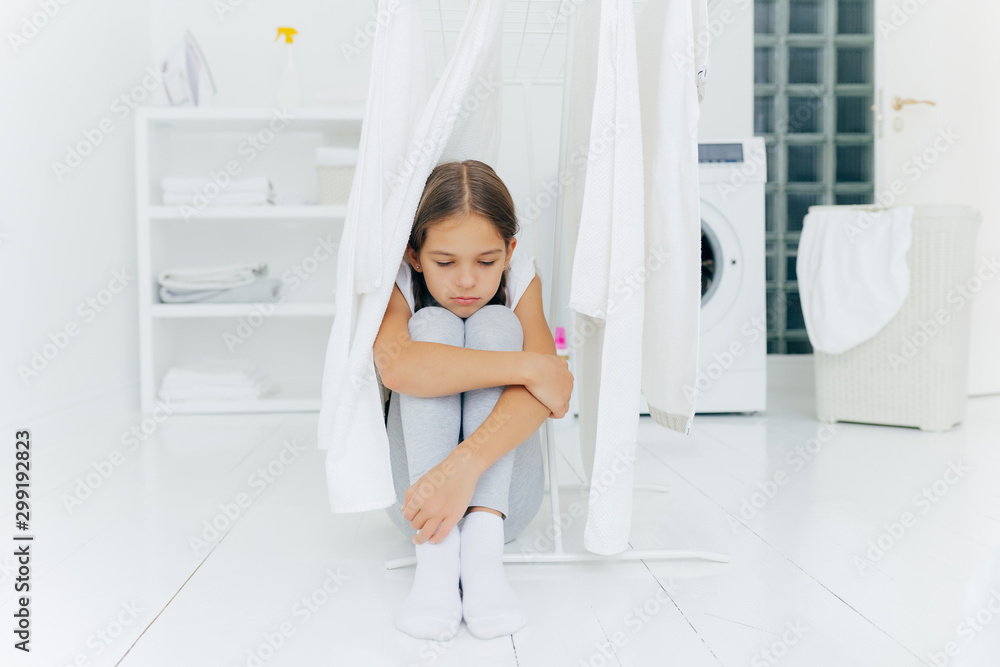 Small adorable girl sits on floor, being punished by parents, poses near clothes dryer, focused down with sad expression, washing machine, basket with laundry and console, thinks over something Small adorable girl sits on floor, being punished by parents, poses near clothes dryer, focused down with sad expression, washing machine, basket with laundry and console, thinks over something
