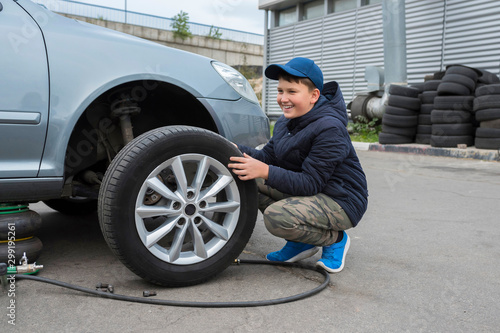 The boy helps at a car service. Replacing the wheels on the car, the jack holds the body in a raised position, with the wheel removed. © Andrii