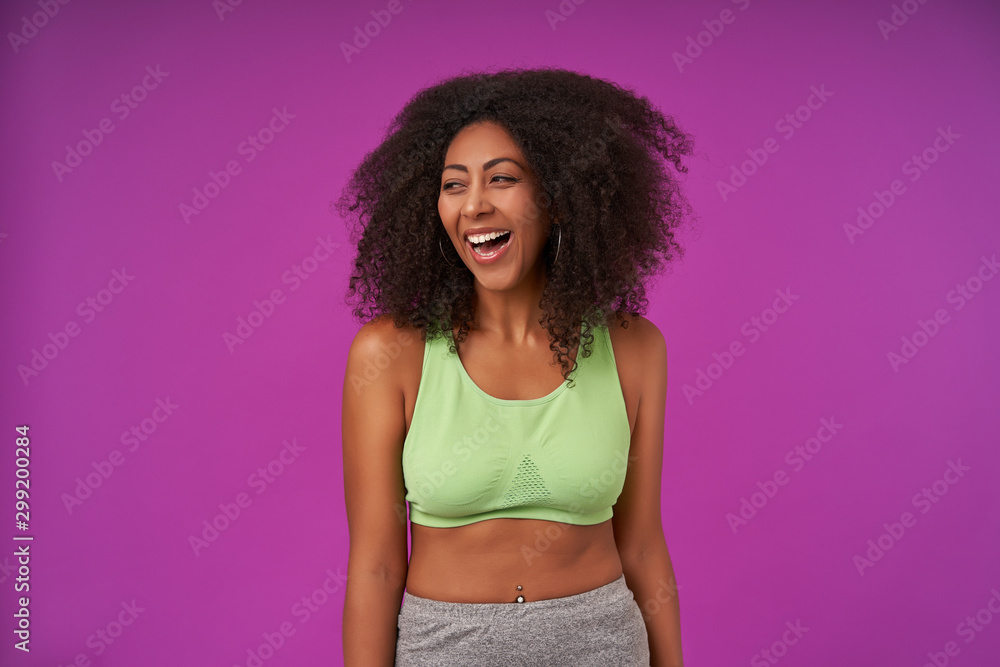 Portrait of beautiful young curly dark skinned woman in casual clothes posing over purple background with hands down, looking aside with joyful and charming smile, demonstrating white perfect teeth Portrait of beautiful young curly dark skinned woman in casual clothes posing over purple background with hands down, looking aside with joyful and charming smile, demonstrating white perfect teeth