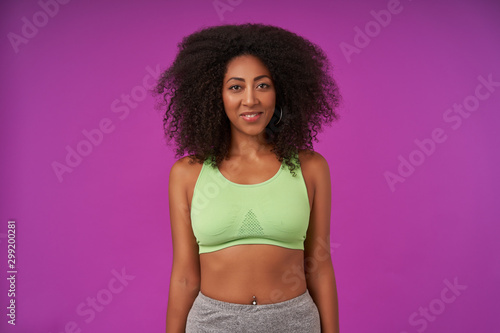 Studio shot of sporty young attractive dark skinned female with curly hair wearing light green top, standing over purple background with hands down, looking at camera with sincere smile