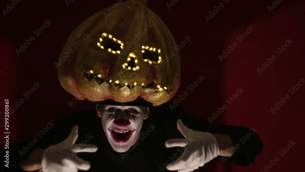 Scary man in the makeup of a clown holds a pumpkin for Halloween and puts it on his head.Terrible clown puts on a mask in the shape of a pumpkin on his head. Terrible clown holds in his hands a Jack-O Scary man in the makeup of a clown holds a pumpkin for Halloween and puts it on his head.Terrible clown puts on a mask in the shape of a pumpkin on his head. Terrible clown holds in his hands a Jack-O