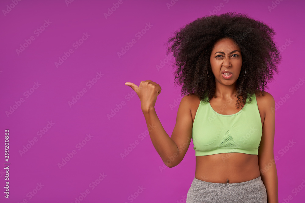 Portrait of sporty young dark skinned woman with curly hair wearing light green top, looking at camera with pout and showing aside with thumb, isolated over purple background Portrait of sporty young dark skinned woman with curly hair wearing light green top, looking at camera with pout and showing aside with thumb, isolated over purple background