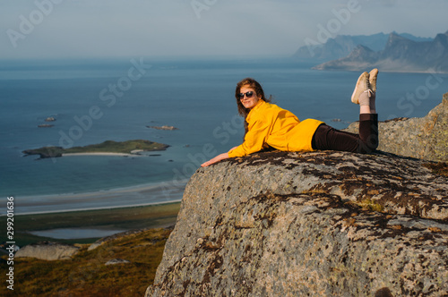 Smiling girl in yellow jacket and sunglasses lies on stone in on the edge of rocks in mountains and looking to camera photo