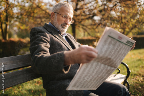 Handsome grandfather with a beautiful beard in a gray jacket sits on a bench in the park and reads a newspaper. Senior gray-haired man in glasses
