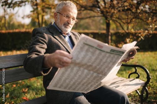 Handsome grandfather with a beautiful beard in a gray jacket sits on a bench in the park and reads a newspaper. Senior gray-haired man in glasses