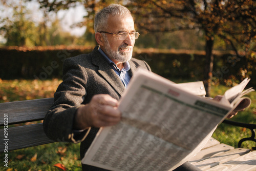 Handsome grandfather with a beautiful beard in a gray jacket sits on a bench in the park and reads a newspaper. Senior gray-haired man in glasses