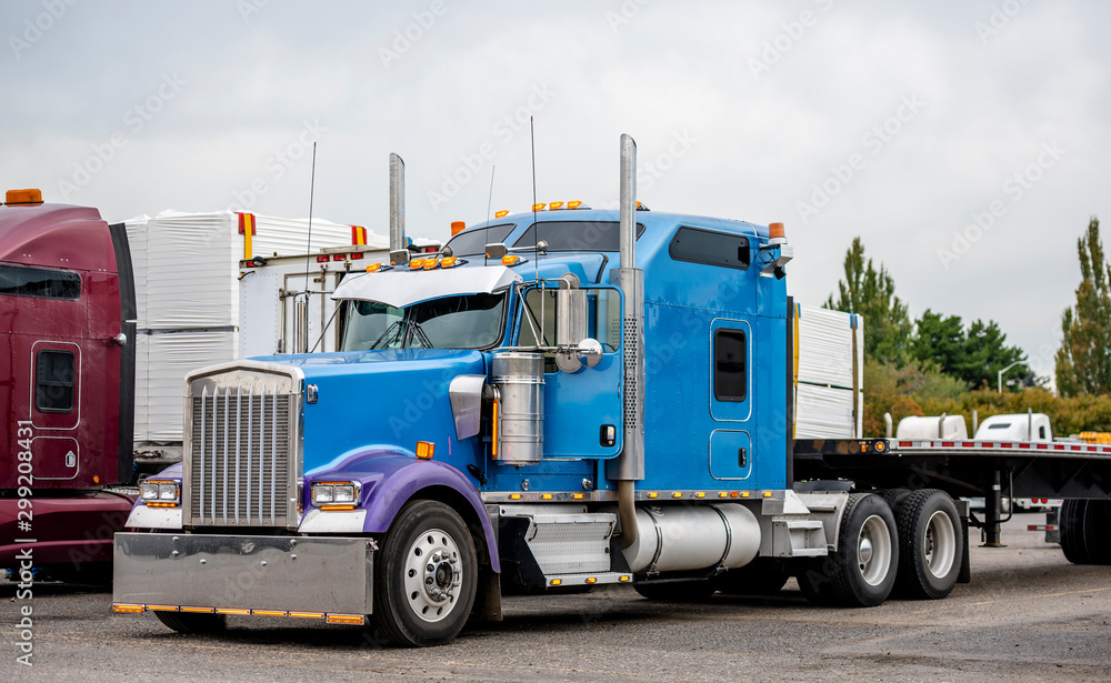 Classic big rig blue semi truck with long sleeping compartment and flat bed semi trailer standing on warehouse parking lot