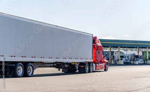 Red big rig semi truck transporting cargo in dry van semi trailer running on the truck stop to fuel station for refuel and continue delivery