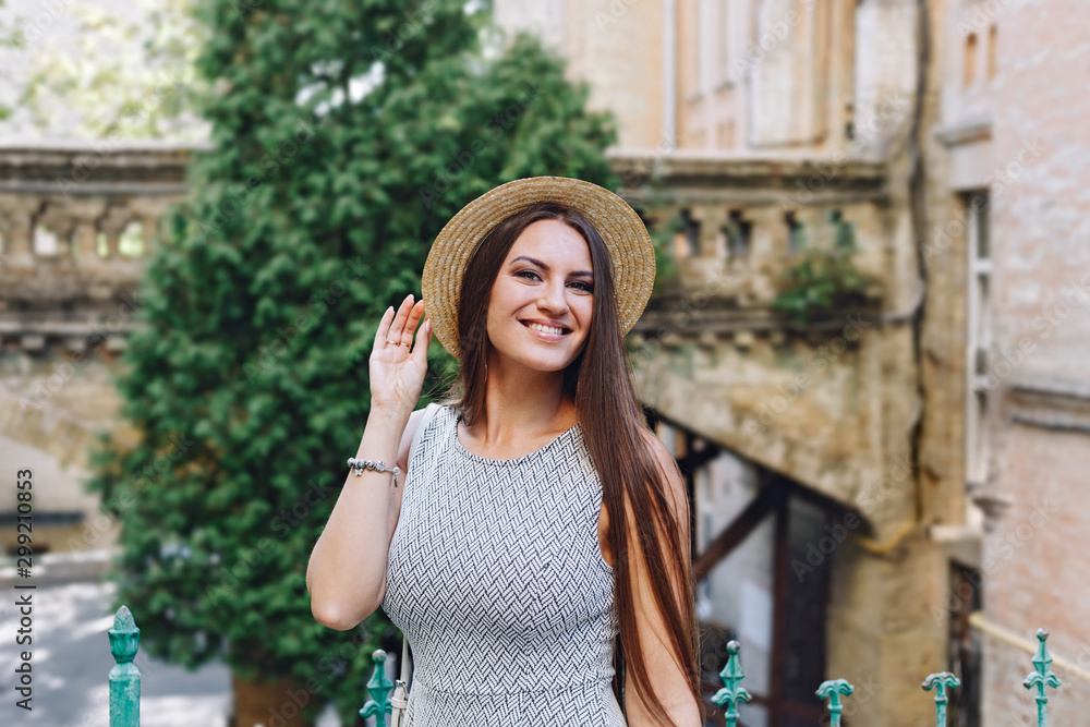 Beautiful brunette girl with long hair in a summer dress and a straw hat in the city on a background of buildings. Smiling attractive girl