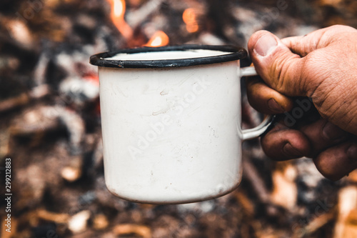 Man hand holds a hot cup of coffee at the background of the campfire. Concept adventure active