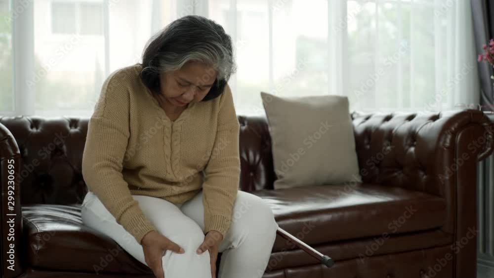 Senior adult with a painful expression from a serious legs and knee pain having difficulty getting up, sitting down on the sofa and moving around the house Senior adult with a painful expression from a serious legs and knee pain having difficulty getting up, sitting down on the sofa and moving around the house