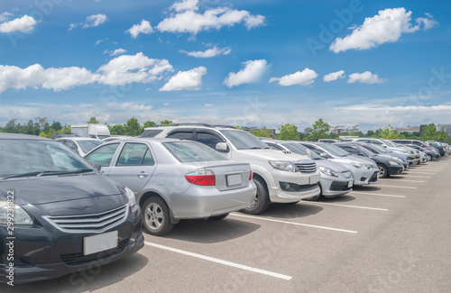Car parking in large asphalt parking lot with trees, white cloud and blue sky background