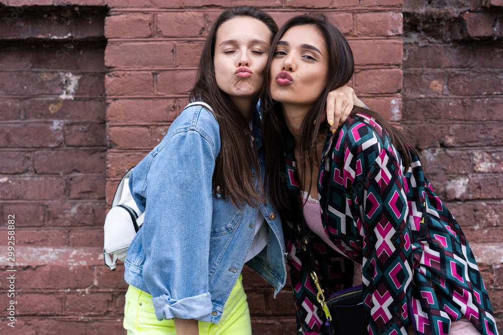 Two young beautiful smiling hipster girls. Positive models having fun. Giving air kiss