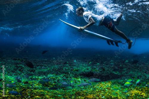 Surfer dives under the wave with his surf board. Underwater shot with vivid coral reef and lots of fish swimming under the surfer