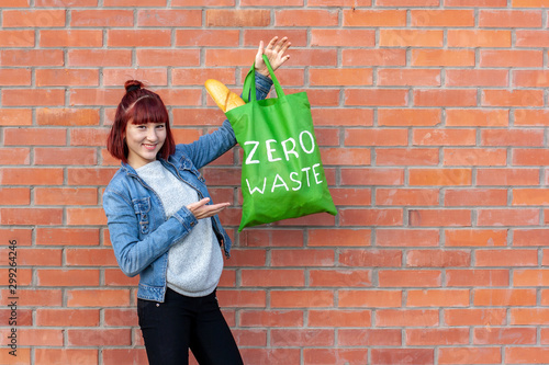 Smiling young girl demonstrates a green textile eco bag with white text zero waste on a brick wall background. The girl has red short hair. French bread in a bag. Copy space on the right.