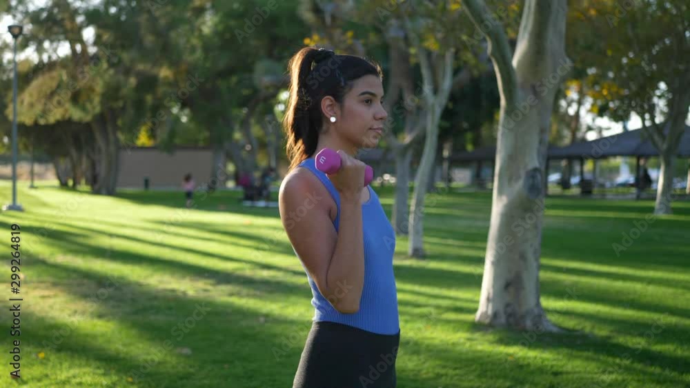 A young woman doing a dumbbell workout in the park performing bicep curls to build arm muscle and strength SLOW MOTION ORBIT LEFT. A young woman doing a dumbbell workout in the park performing bicep curls to build arm muscle and strength SLOW MOTION ORBIT LEFT.