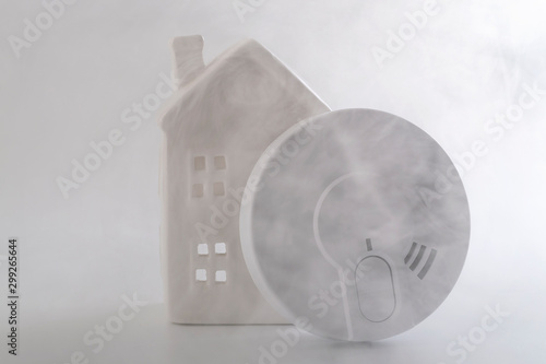 Fire prevention, home security and monitoring dangerous fumes conceptual idea with generic smoke detector and miniature house surrounded by thick smoke isolated on white background