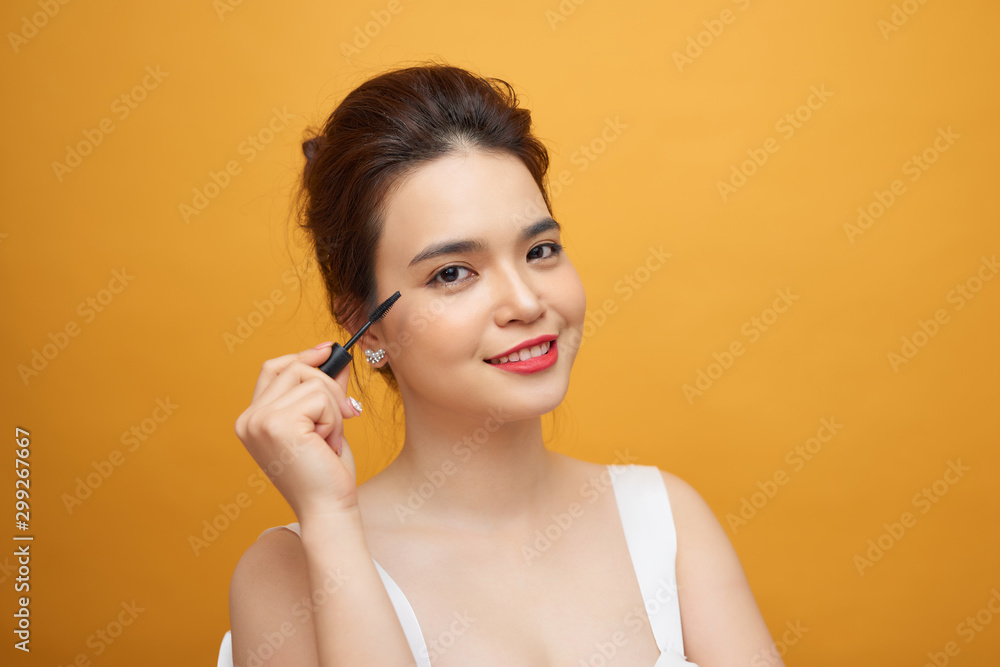 Closeup portrait of attractive young woman putting some mascara onto her eyelashes with make up brush over yellow background Closeup portrait of attractive young woman putting some mascara onto her eyelashes with make up brush over yellow background