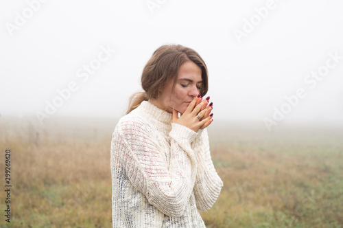 Girl closed her eyes, praying in a field during beautiful fog. Hands folded in prayer concept for faith