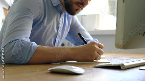 A man in a blue shirt makes a note in a notebook. Close-up: the man bends over the desk and makes notes with a pen, leaning on the table.