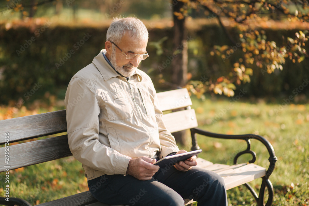Fototapeta premium Handsome elderly man sitting on the bench and use tablet for scrolling in internet. Background of autumn tree