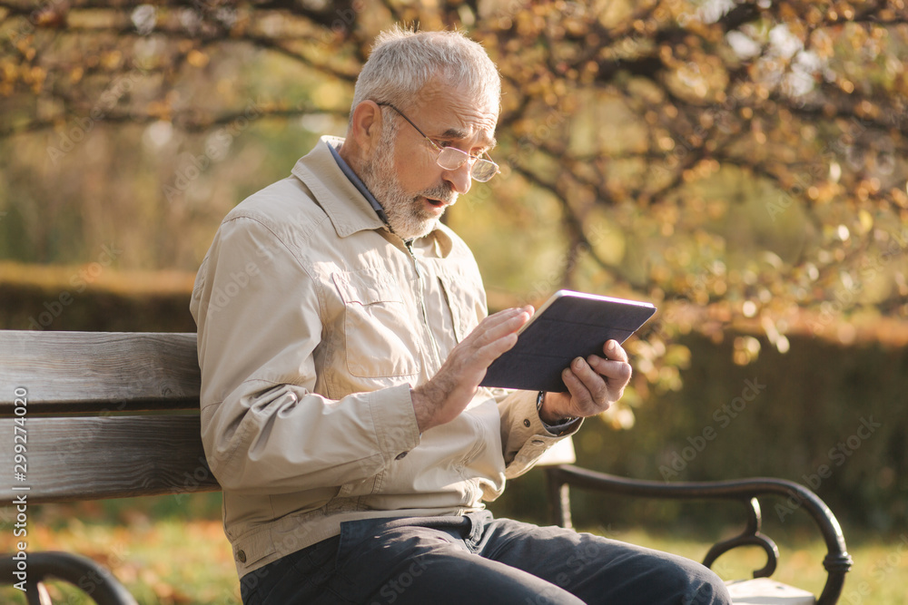 Handsome elderly man sitting on the bench and use tablet for scrolling in internet. Background of autumn tree Handsome elderly man sitting on the bench and use tablet for scrolling in internet. Background of autumn tree