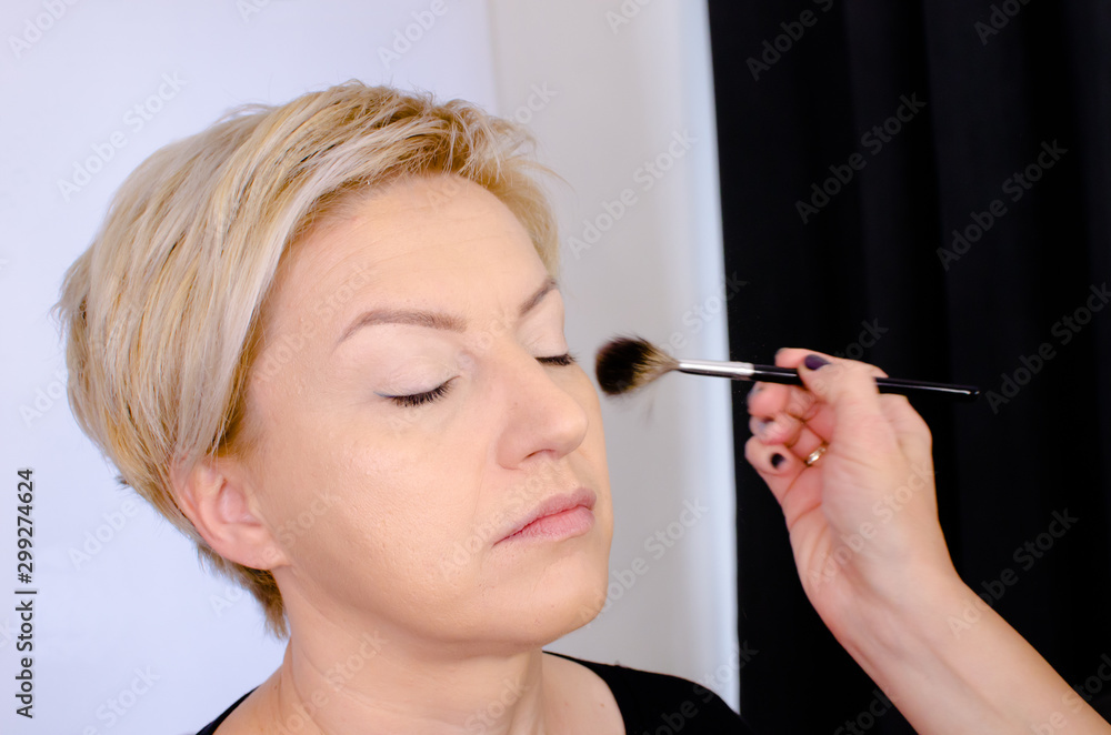 Fototapeta premium Make-up artist working with brush on model face before applying make up Portrait of senior blonde woman in beauty studio with closed eyes. Close up of putting powder with cosmetic brush