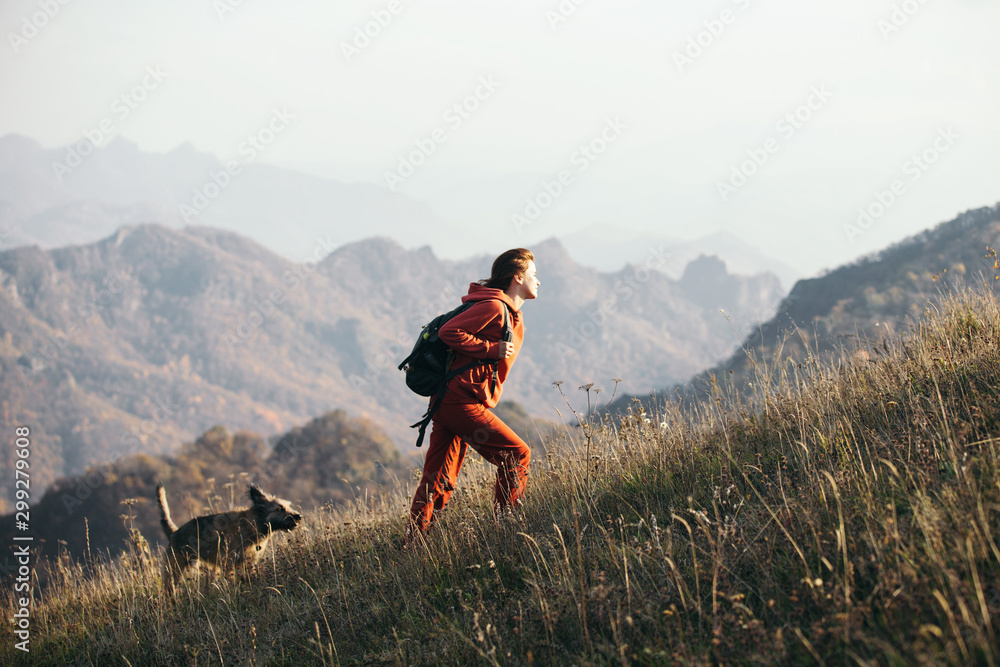 Beautiful woman traveler climbs uphill with a dog on a background of mountain views. She is with a backpack and in red clothes. - obrazy, fototapety, plakaty 