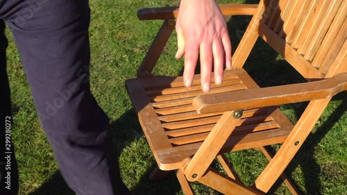 Slow motion close POV shot of a man wearing a blue fleece pullover in the bright cool sunshine, unfolding a wooden garden / yard chair, then sitting down and leaning back in it to relax.