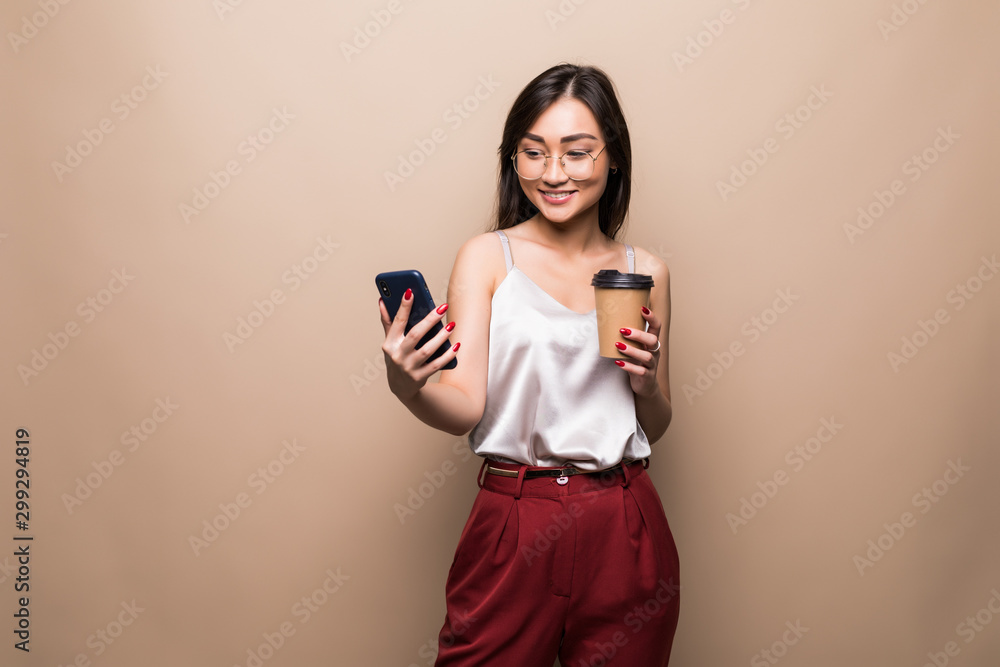 Full length portrait of a smiling asian woman using mobile phone while holding cup of coffee to go isolated over beige background Full length portrait of a smiling asian woman using mobile phone while holding cup of coffee to go isolated over beige background